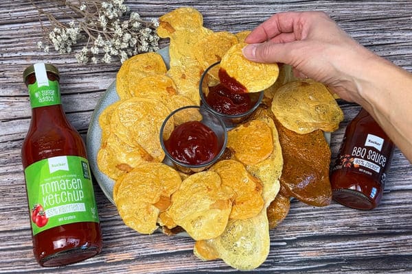 Selbst gemachte Protein Chips aus Harzer Käse auf einem Teller mit zuckerfreien Saucen in zwei keinen Schälchen in der Mitte des Tellers und je einer Flasche Ketchup und BBQ Sauche von Xucker